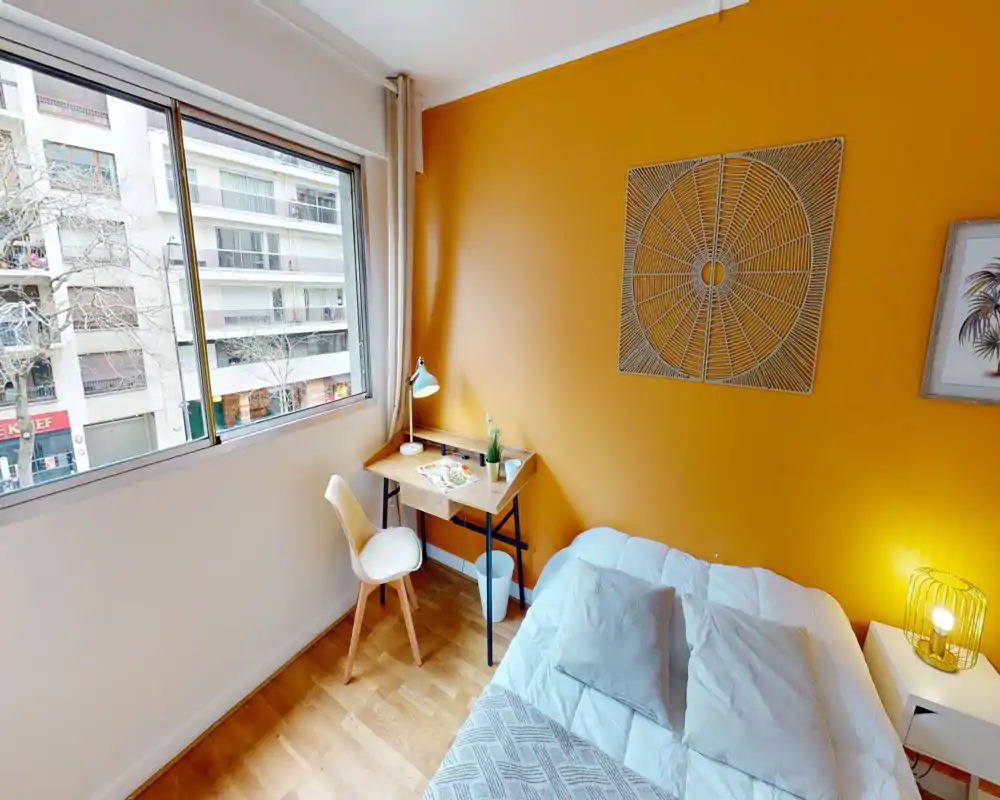 Chambre compacte et lumineuse avec un lit double confortable, mur accent orange et un petit bureau près d'une grande fenêtre apportant beaucoup de lumière naturelle — chaleureuse et bien décorée.