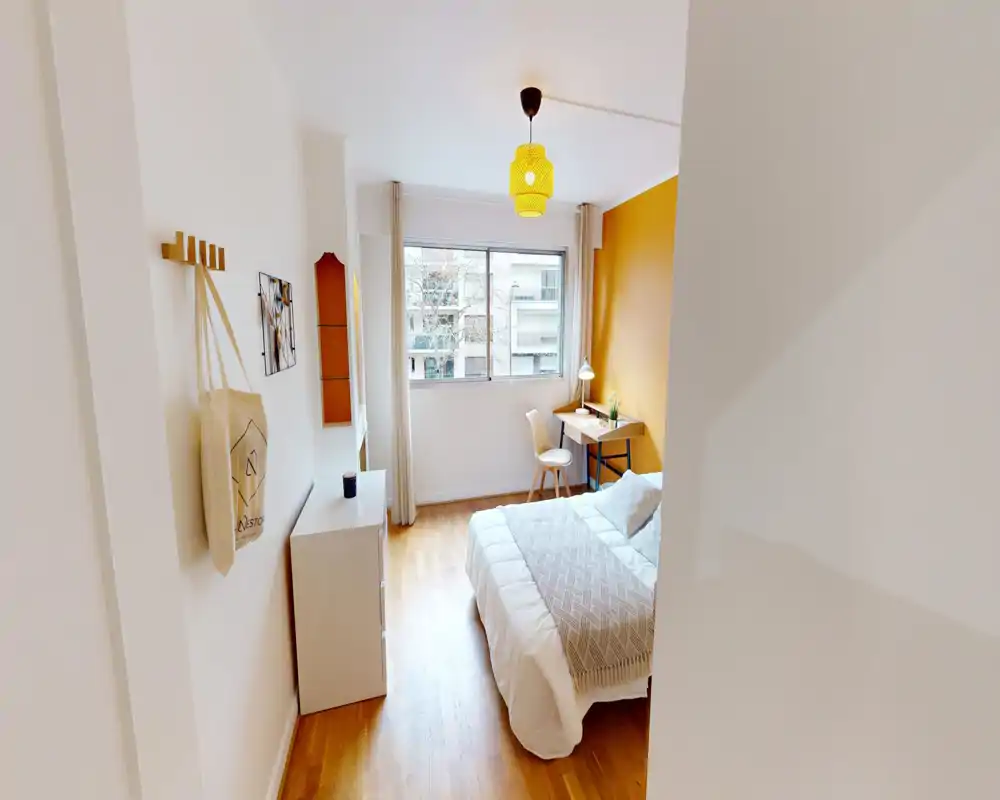 Chambre lumineuse et bien mise en scène avec un lit double confortable, un mur d'accent chaud et un bureau près de la fenêtre — parfaite pour le repos et le travail.