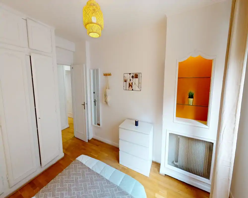 Chambre lumineuse et soignée avec lit partiellement visible au premier plan, placards encastrés blancs, commode et suspension chaude apportant une ambiance cosy.