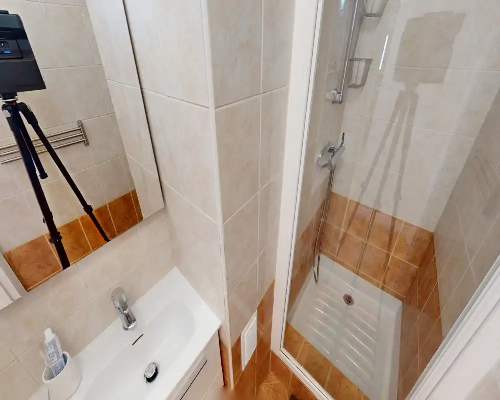 Compact, well-lit bathroom with a corner shower cabin and a small sink with mirrored cabinet. Clean tiled walls in neutral tones and modern chrome fixtures provide a neat, functional space ideal for short-stay rentals.