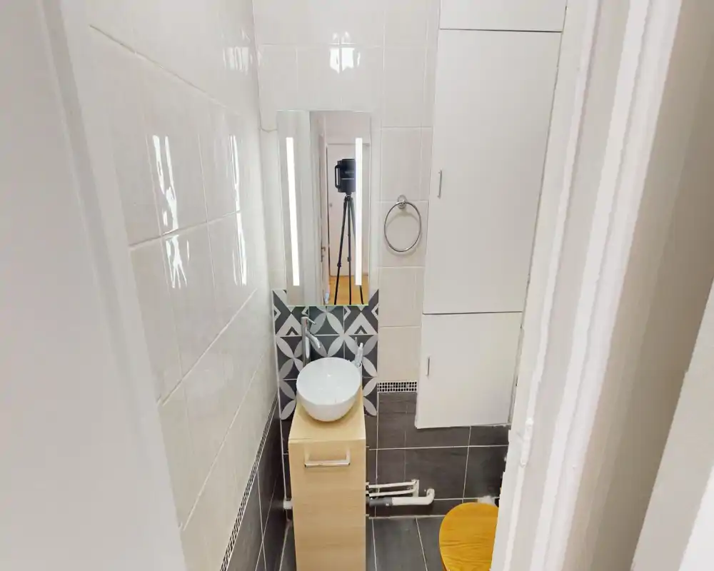Compact modern bathroom with tiled walls, a countertop vessel sink on a small wooden vanity, lit mirror and a visible toilet seat. The space is well lit and neatly finished.