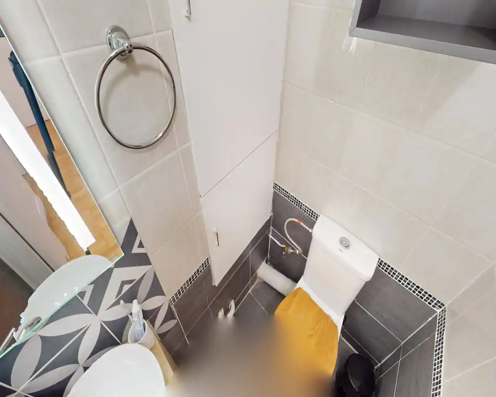 Compact tiled bathroom with a close-up view of a toilet with wooden seat, sink countertop and mirror; modern mosaic trim and neutral tiles create a clean, functional space.
