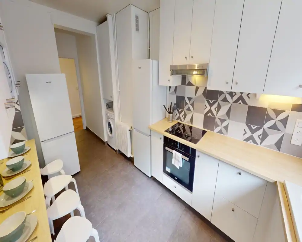 Modern galley kitchen with white cabinets, patterned tile backsplash, electric cooktop and integrated oven. Light wood countertops and a breakfast bar with stools create a clean, functional space.