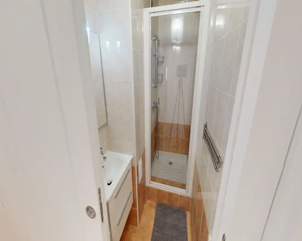 Compact, fully tiled bathroom with a standing shower, small sink and mirror — bright and neatly presented, ideal for a small apartment.