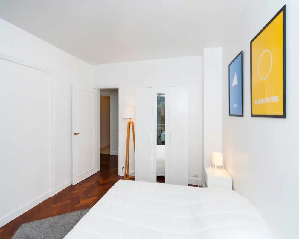 Bright attic bedroom with slanted white ceilings, visible bed with white bedding and framed prints on the wall. The room feels airy and simply decorated, suitable for a cozy single or couple.