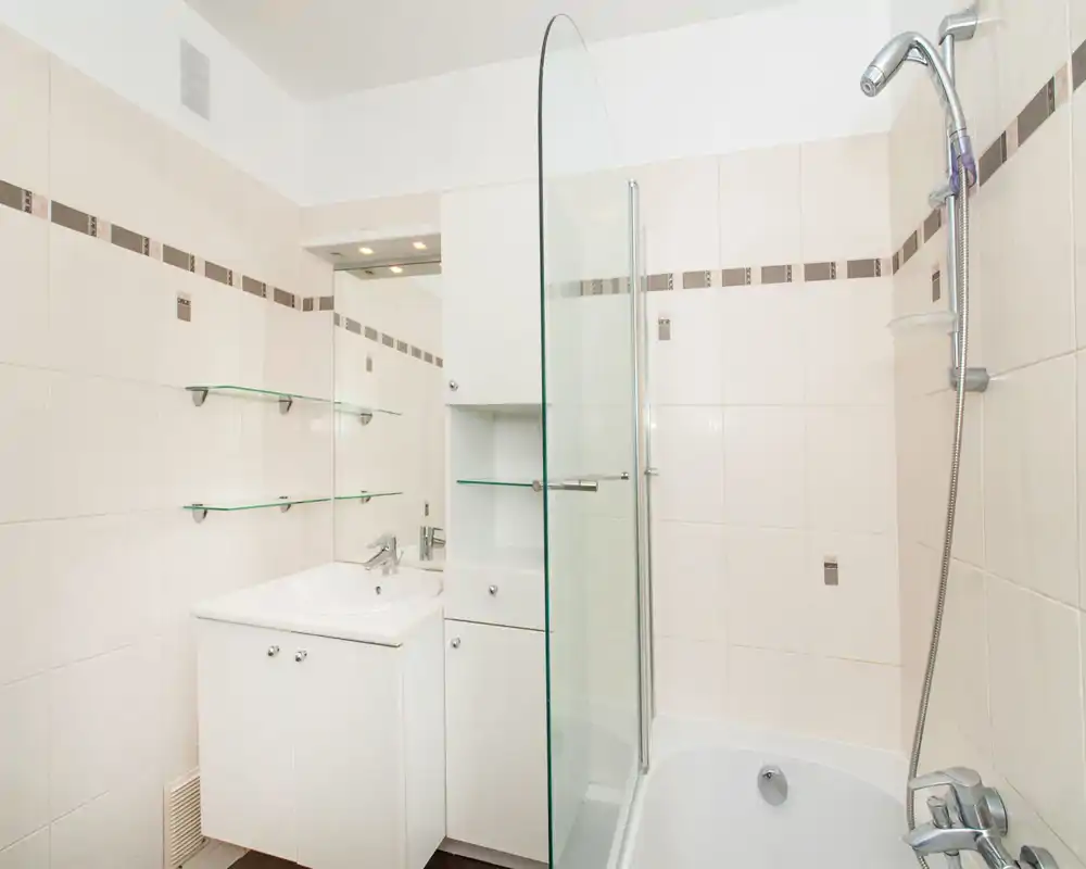 Clean, modern bathroom with a bathtub, shower head, glass shower screen and a white sink cabinet; pale tiles and glass shelves create a bright, minimalist look.