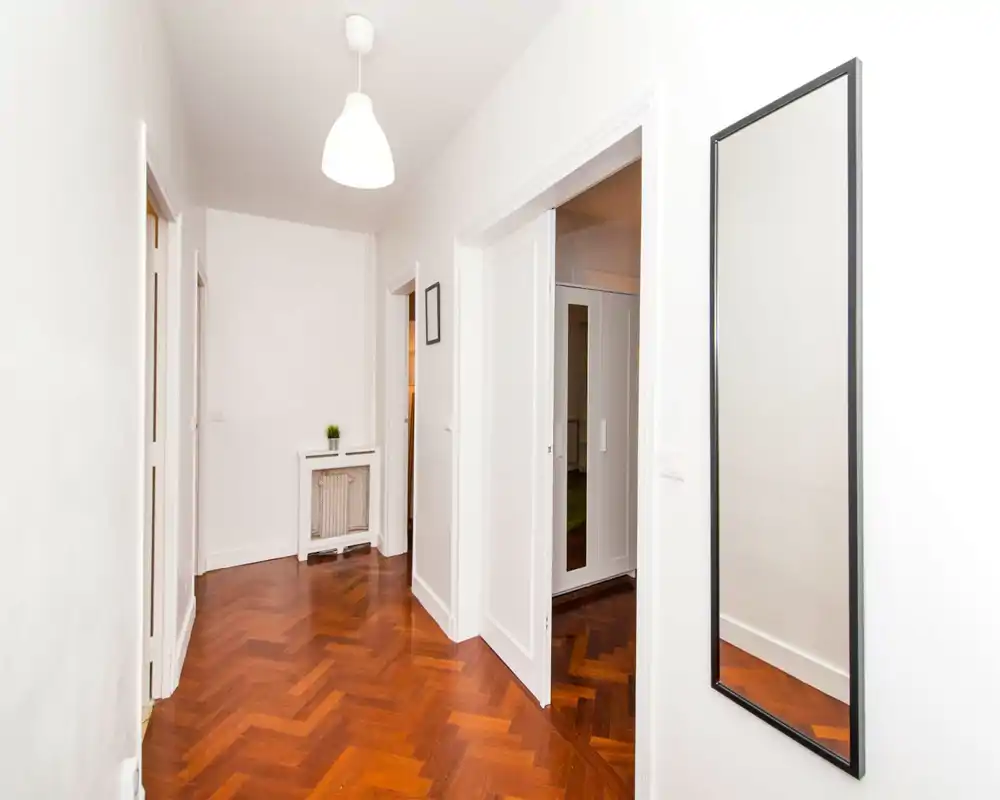 Bright, minimal hallway with herringbone wood floor, white walls, a wall mirror and pendant light — a clean and welcoming entrance area.
