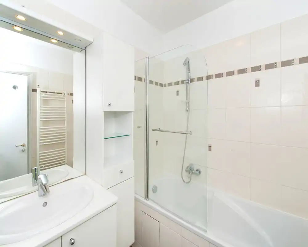 Clean, modern bathroom with a bathtub and glass shower screen, white vanity with sink and a mirrored cabinet. Bright neutral tiles and chrome fixtures create a fresh, minimalist look.
