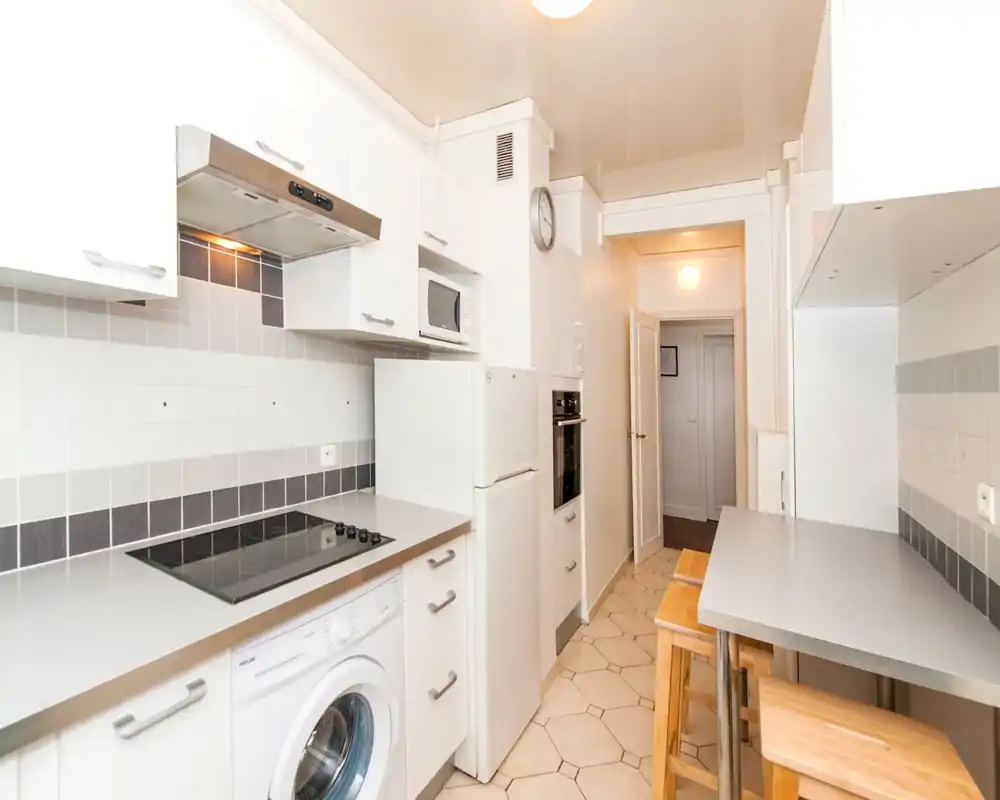 Bright, compact modern kitchen with white cabinetry, electric cooktop, built-in microwave and fridge, plus a small breakfast bar with wooden stools.