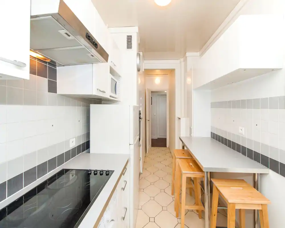 Bright, narrow modern kitchen with a tiled backsplash, induction hob, built-in appliances and a breakfast counter with wooden stools — ideal for compact urban apartments.