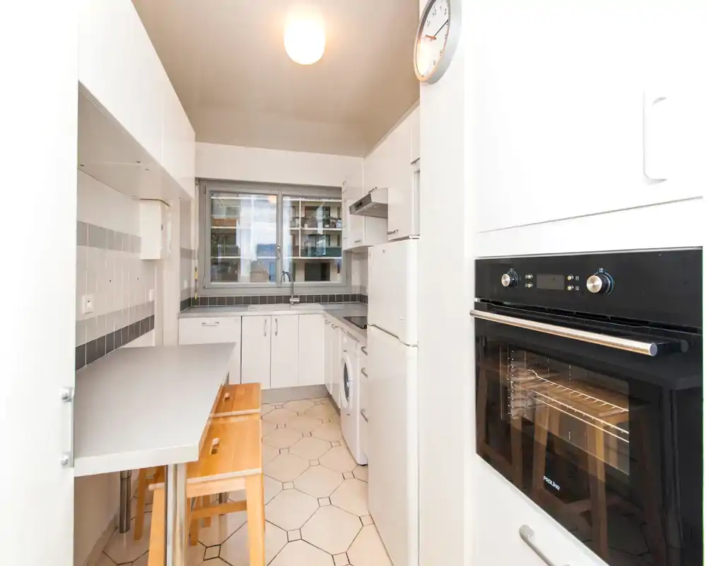 Bright, compact modern kitchen with white cabinets, built-in oven, refrigerator and a small breakfast counter with wooden stools — ideal for a city apartment.