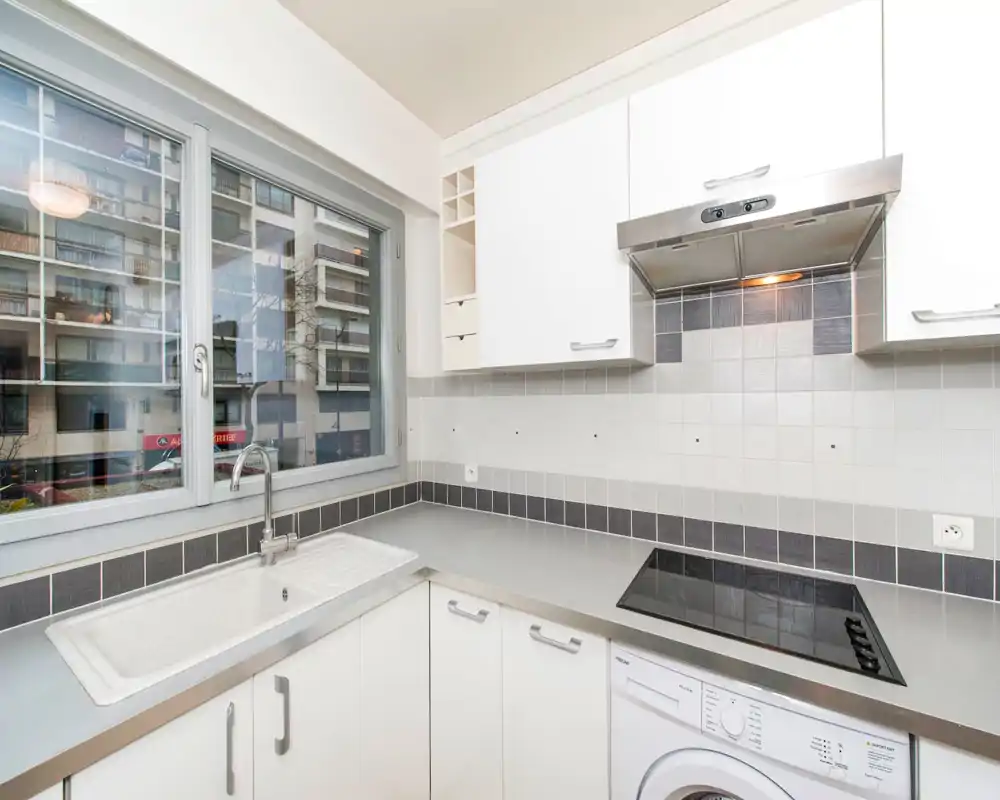 Bright modern kitchen with a large window, stainless-steel countertop, built-in cooktop and sink — clean, functional space ideal for a compact apartment.