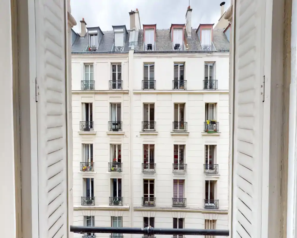 Open window with shutters framing a view of a classic Parisian building façade and small balconies — ideal to highlight the apartment’s street-facing aspect and natural light.