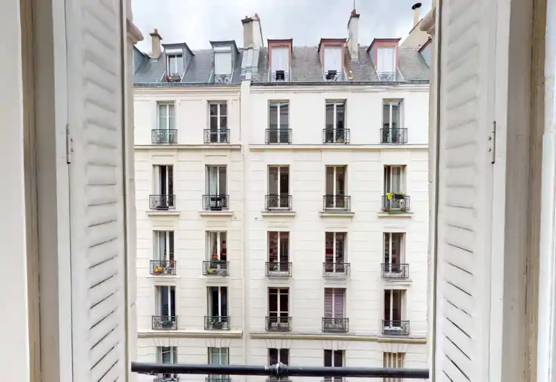 Open window with shutters framing a view of a classic Parisian building façade and small balconies — ideal to highlight the apartment’s street-facing aspect and natural light.