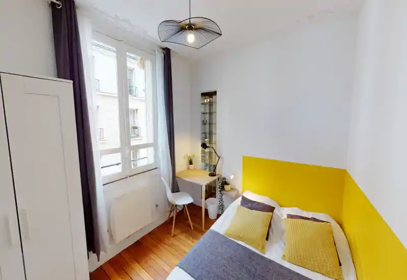 Bright, well-styled bedroom with a large window, cozy textiles and a yellow accent headboard. A compact desk and wardrobe make it practical for a single occupant.