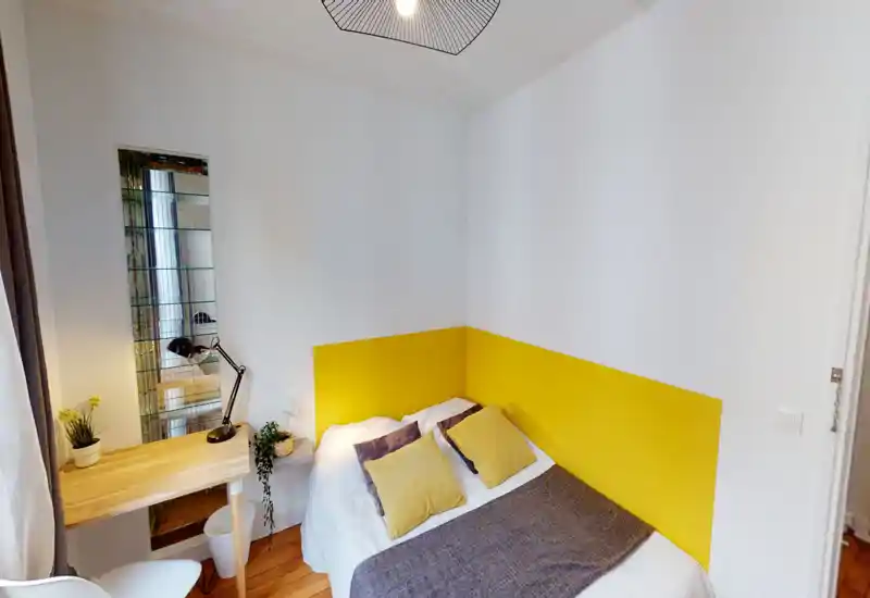 Cozy small bedroom with a double bed against a cheerful yellow accent wall, textured throw and cushions, a compact wooden desk with lamp, and bright white walls that enhance natural light.