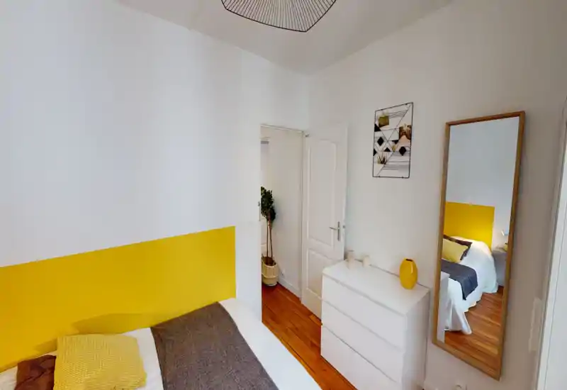 Bright, modern bedroom with a yellow accent headboard, wooden floor and a full-length mirror reflecting the bed. The room feels fresh and styled with minimal, cozy textiles and decorative accents.