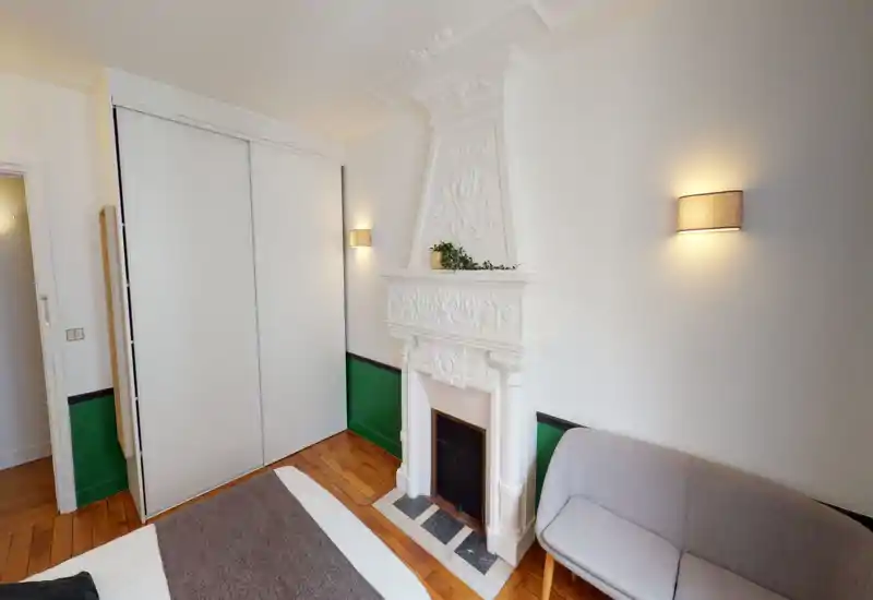 Bright, stylish bedroom featuring a decorative white fireplace, built-in sliding wardrobe and a comfortable bed with warm wall lighting—elegant hardwood floors and touches of greenery add charm.