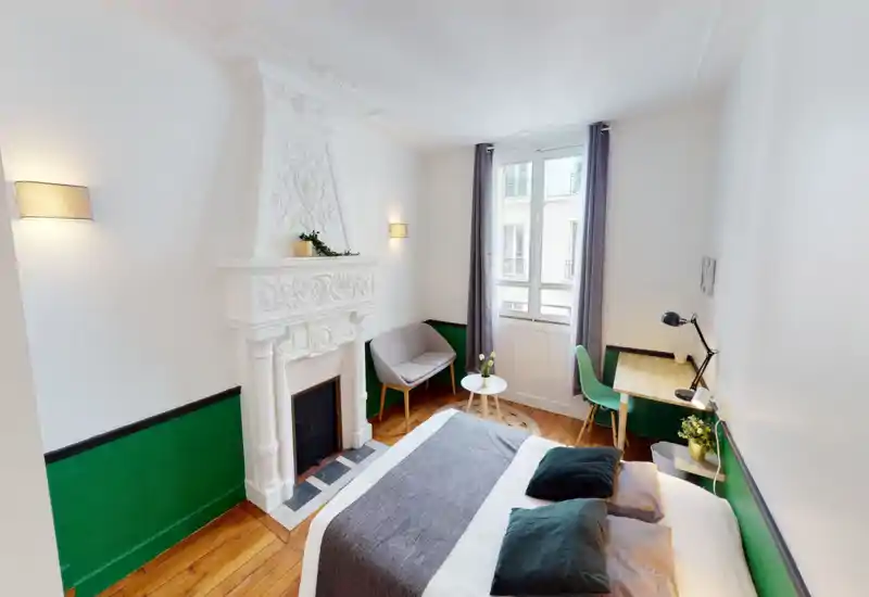 Bright, stylish bedroom with a comfortable double bed, decorative fireplace, small seating area and a work desk near the window — ideal for a cozy city stay.
