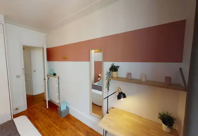 Angolo camera ben arredato con scrivania in legno, mensola sospesa e specchio alto che riflette il letto; pavimento in legno caldo e luce soffusa che creano un’atmosfera accogliente e luminosa.