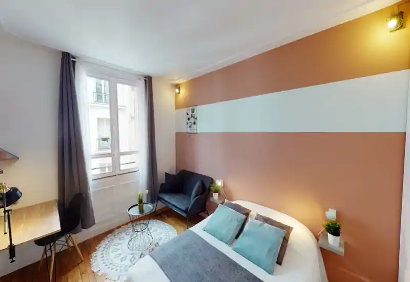 Camera da letto luminosa e di stile con letto matrimoniale, tessili morbidi e parete d'accento calda. Luce naturale da una finestra alta, piccolo angolo salotto e scrivania compatta rendono la stanza funzionale e accogliente.