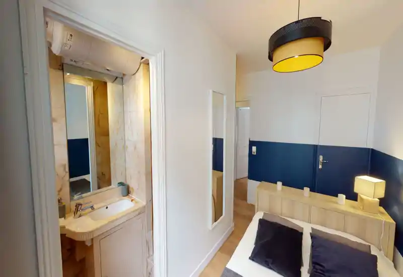 Bright, neatly styled bedroom with a wooden headboard, decorative pillows and a bedside lamp; an ensuite sink/bathroom is visible through a doorway. The room features a two-tone wall (white and deep blue) and warm wooden flooring.