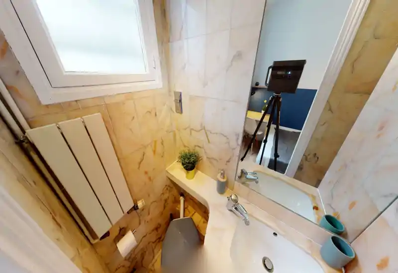 Compact marble-tiled bathroom with a sink, mirror, radiator and toilet; bright natural light from a frosted window and modern chrome fixtures.