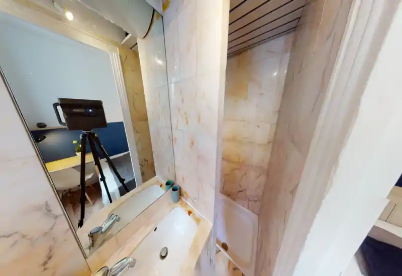 Compact bathroom with marble-look tiled walls, a small shower tray and a sink with a mirror. The photo shows clean fixtures and a bright, well-lit space.