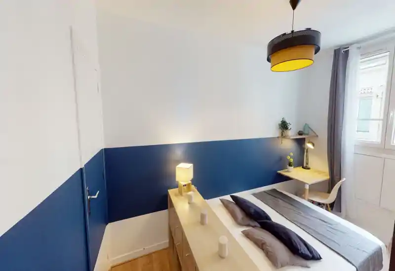 Bright, modern bedroom with a queen-size bed dressed in neutral linens, bedside storage and a small desk by the window. The room features a two-tone blue accent wall, pendant lighting and a roomy window that brings natural light.