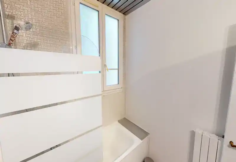 Bright, compact bathroom with a bathtub and a frosted window; partial frosted glass shower screen and visible handheld shower head.