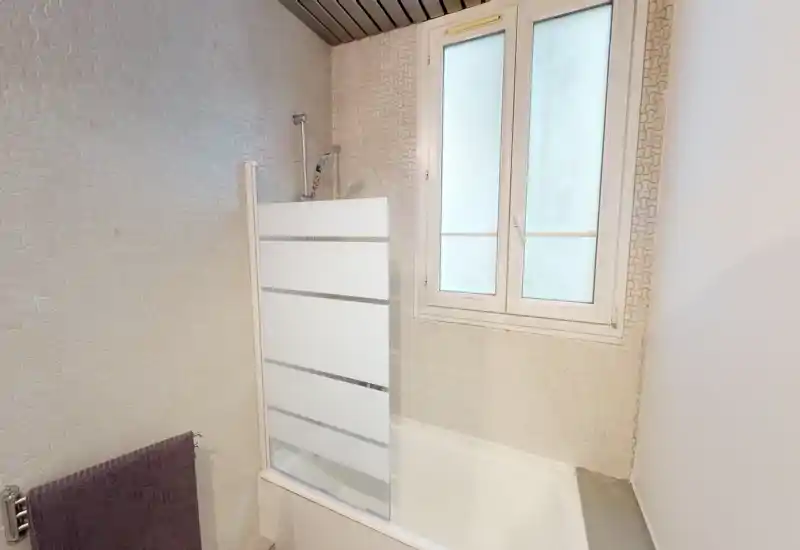 Bright, compact bathroom with a bathtub, frosted window and a half-glass shower screen. Neutral patterned walls and a towel rail give a tidy, functional impression.