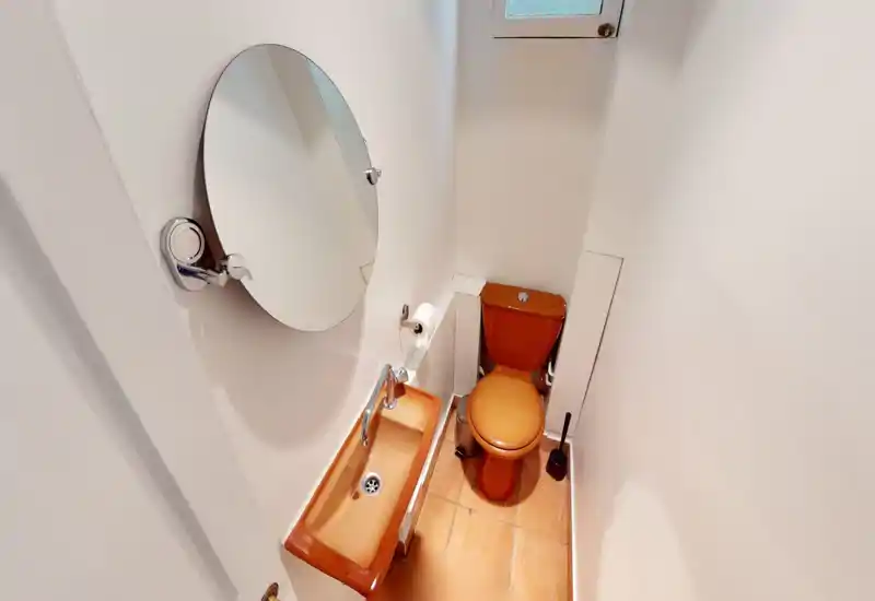 Compact bathroom with a small orange toilet and matching narrow sink, round wall mirror and tiled floor; bright white walls make the fixtures stand out.