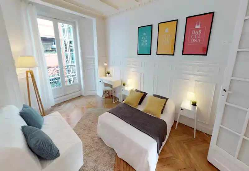 Bright, stylish bedroom with a double bed, bedside tables and a small workspace. Large window and wooden herringbone floor create an airy Parisian feel.