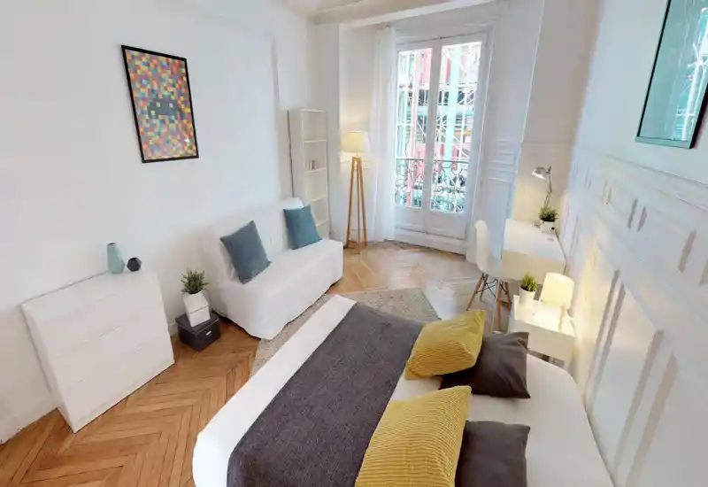 Bright, stylish bedroom with a large visible bed, layered textiles and a balcony window bringing abundant natural light. The room features a small sofa, desk area and tasteful decor perfect for a rental listing.