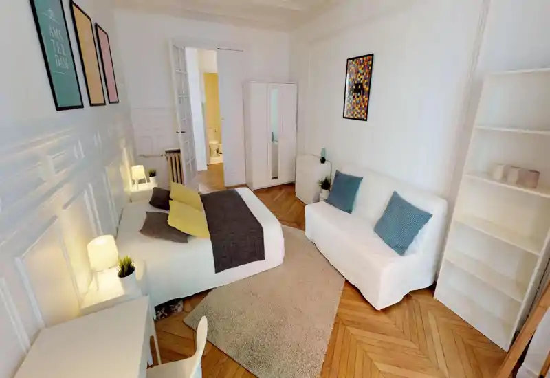 Bright, stylish bedroom with a full double bed centered on a soft rug, bedside lamps, a white sofa and wardrobe — ideal for rental listings highlighting comfort and tasteful décor.