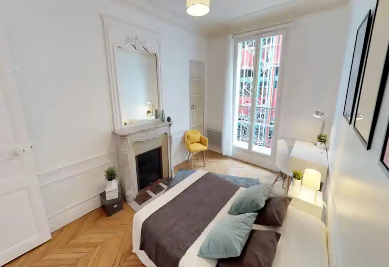 Bright, elegant bedroom with a double bed centered in the foreground, decorative fireplace and large window letting in abundant natural light. Contemporary minimal decor with soft textiles, a cozy rug and a small desk by the window.