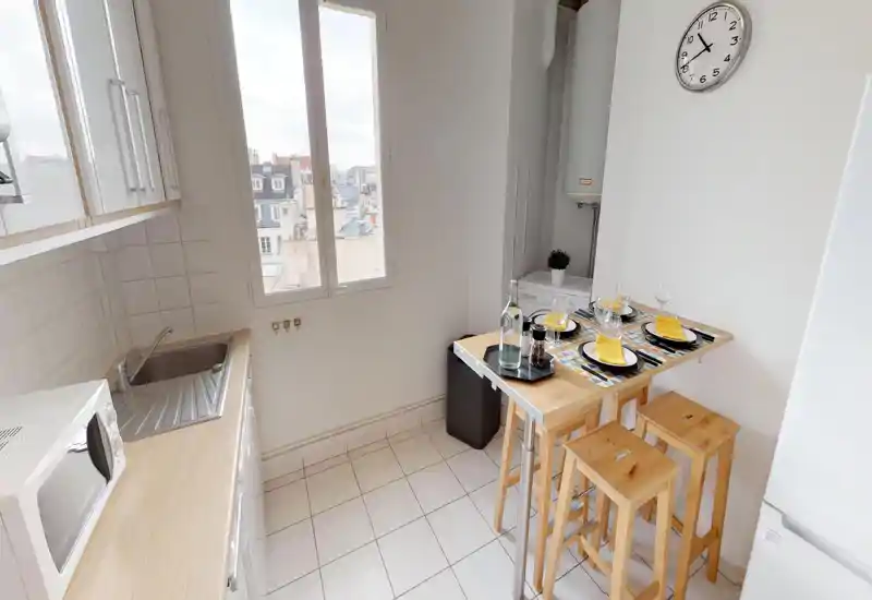 Compact, bright kitchen with a sink, microwave and a small breakfast bar set for four. Large window brings natural light and the wooden stools and table create a warm, functional space ideal for city apartments.