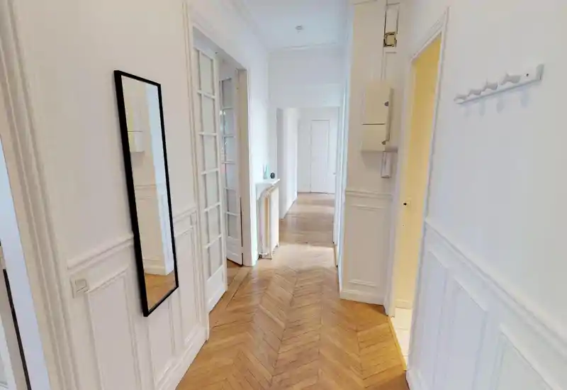 Bright entrance hallway with chevron wood floors, white paneled walls and a full-length mirror — a welcoming, well-lit entry space.