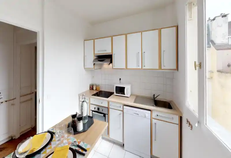 Compact, well-lit kitchen with white cabinets, integrated appliances and a small breakfast counter set for two — bright, tidy and practical for city living.