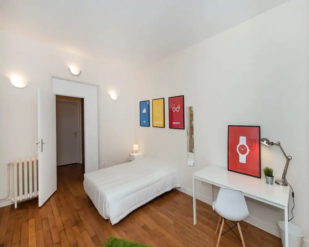 Camera da letto minimal e luminosa con un letto singolo ben rifatto, pareti bianche e pavimento in legno; una scrivania con lampada e quadri crea uno spazio ordinato e funzionale.