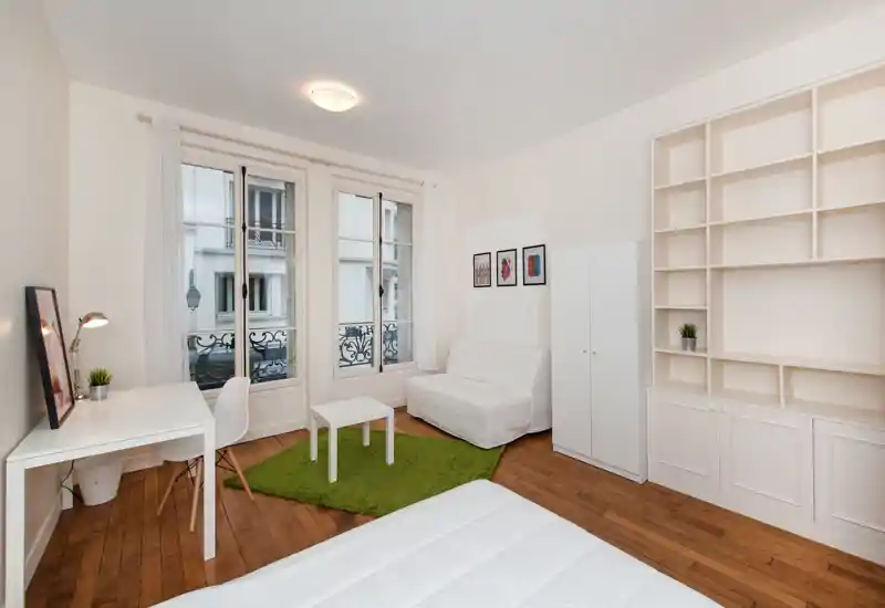 Camera da letto luminosa e minimal con ampie finestre in stile francese, letto bianco in primo piano, piccolo angolo salotto e scaffalature a muro. Pavimento in legno e tappeto verde danno calore.