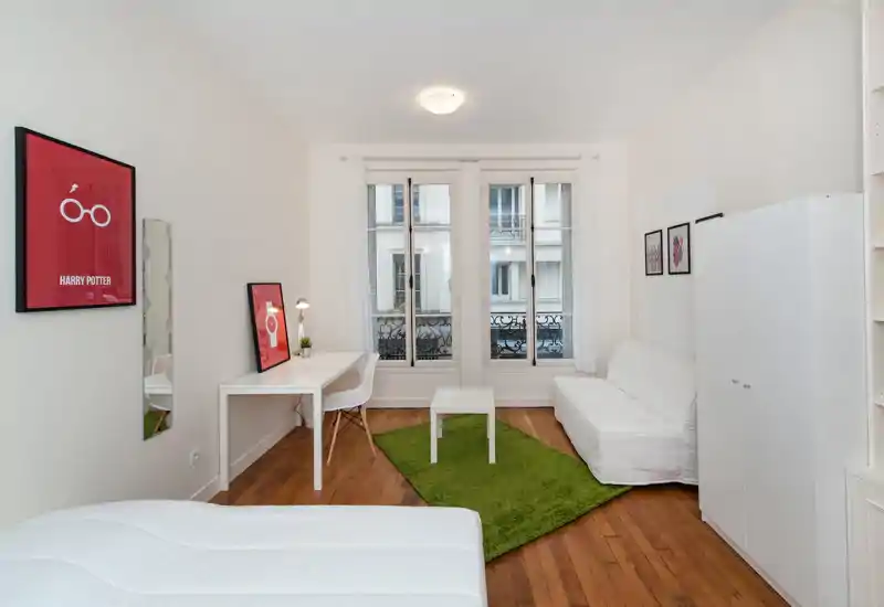 Camera da letto luminosa e ben arredata con letto visibile in primo piano, mobili bianchi, scrivania vicino alla finestra e un accogliente tappeto verde su pavimento in legno.