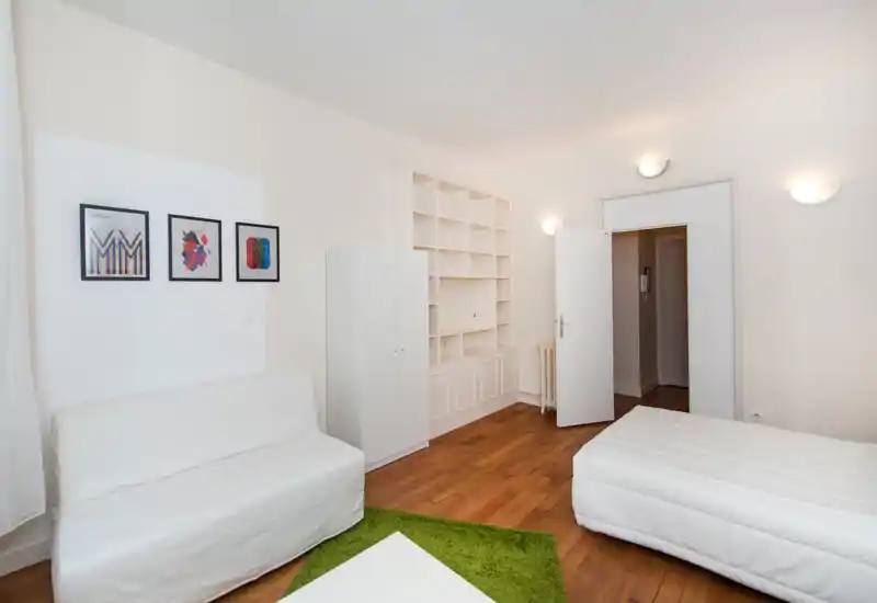 Camera da letto luminosa e moderna con letto e divano bianchi, scaffalatura a muro e pavimento in legno caldo. Arredamento minimal con quadri incorniciati e un tappeto verde che aggiunge un tocco accogliente.