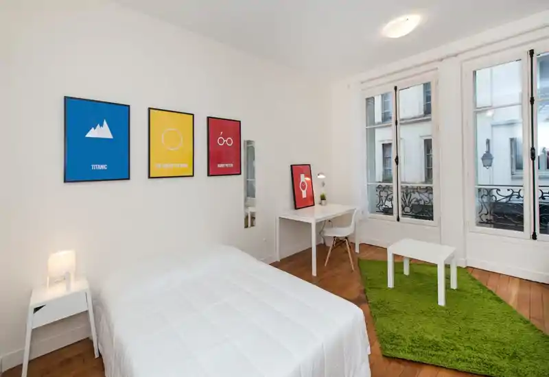 Camera da letto luminosa e moderna con letto matrimoniale ben rifatto, arredi bianchi minimal, stampe colorate alle pareti e grandi finestre che illuminano l’ambiente.