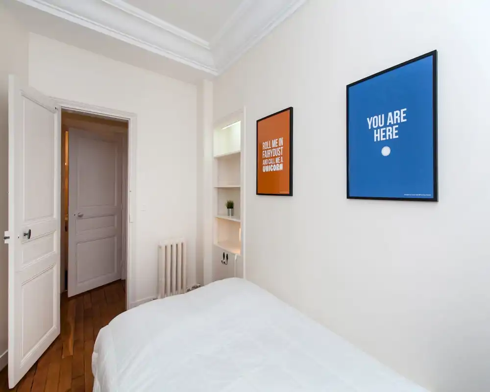 Coin de chambre mansardée lumineux avec vue partielle sur un lit blanc et des cadres décoratifs sous une toiture inclinée — style minimal et moderne, adapté pour une annonce.