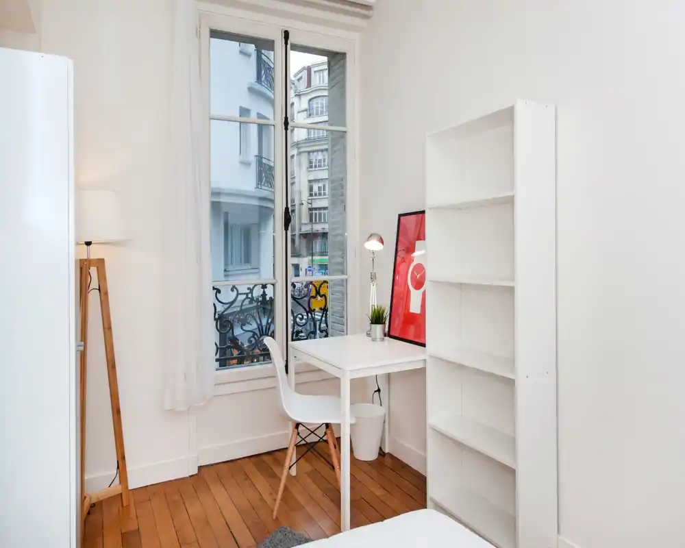 Coin chambre lumineux et minimaliste avec un bureau blanc près d'une grande fenêtre, une chaise simple, des étagères et une partie du lit visible. L'espace est propre, bien éclairé et décoré d'une lampe, d'un cadre et d'une petite plante.