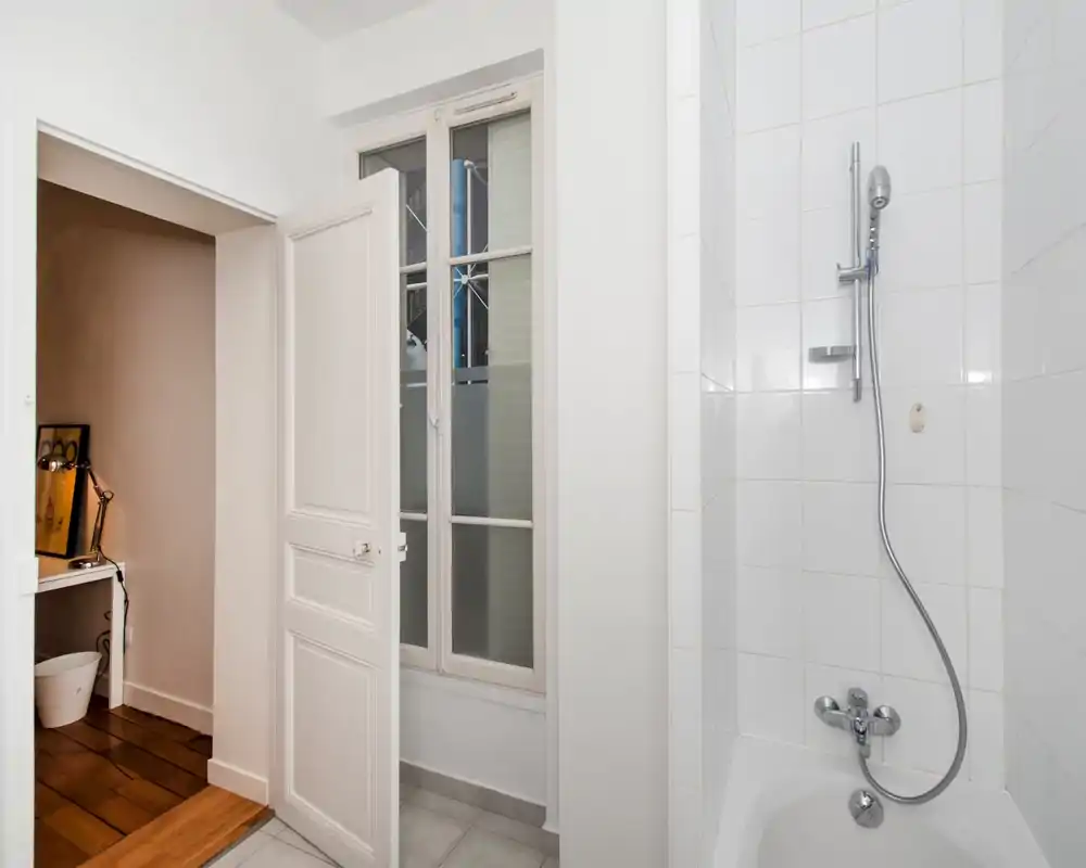 Bagno pulito e semplice con vasca/doccia piastrellata, doccetta a mano e porta che si apre su una stanza adiacente. Piastrelle bianche e rubinetteria cromata creano un aspetto ordinato e minimal.