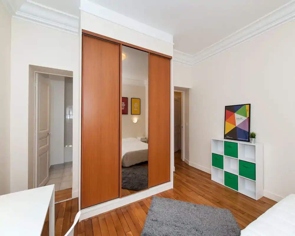 Camera da letto luminosa e semplice con armadio scorrevole in legno e specchio centrale, letto con biancheria bianca, tappeto grigio e mensola minimal con contenitori verdi.