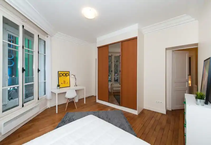 Camera da letto luminosa e ben arredata con grandi finestre, letto parzialmente visibile in primo piano, una scrivania vicino alla finestra e un armadio con specchio che dona profondità.