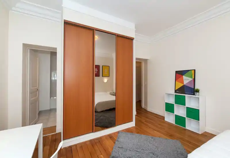 Camera da letto luminosa e semplice con armadio scorrevole in legno e specchio centrale, letto con biancheria bianca, tappeto grigio e mensola minimal con contenitori verdi.